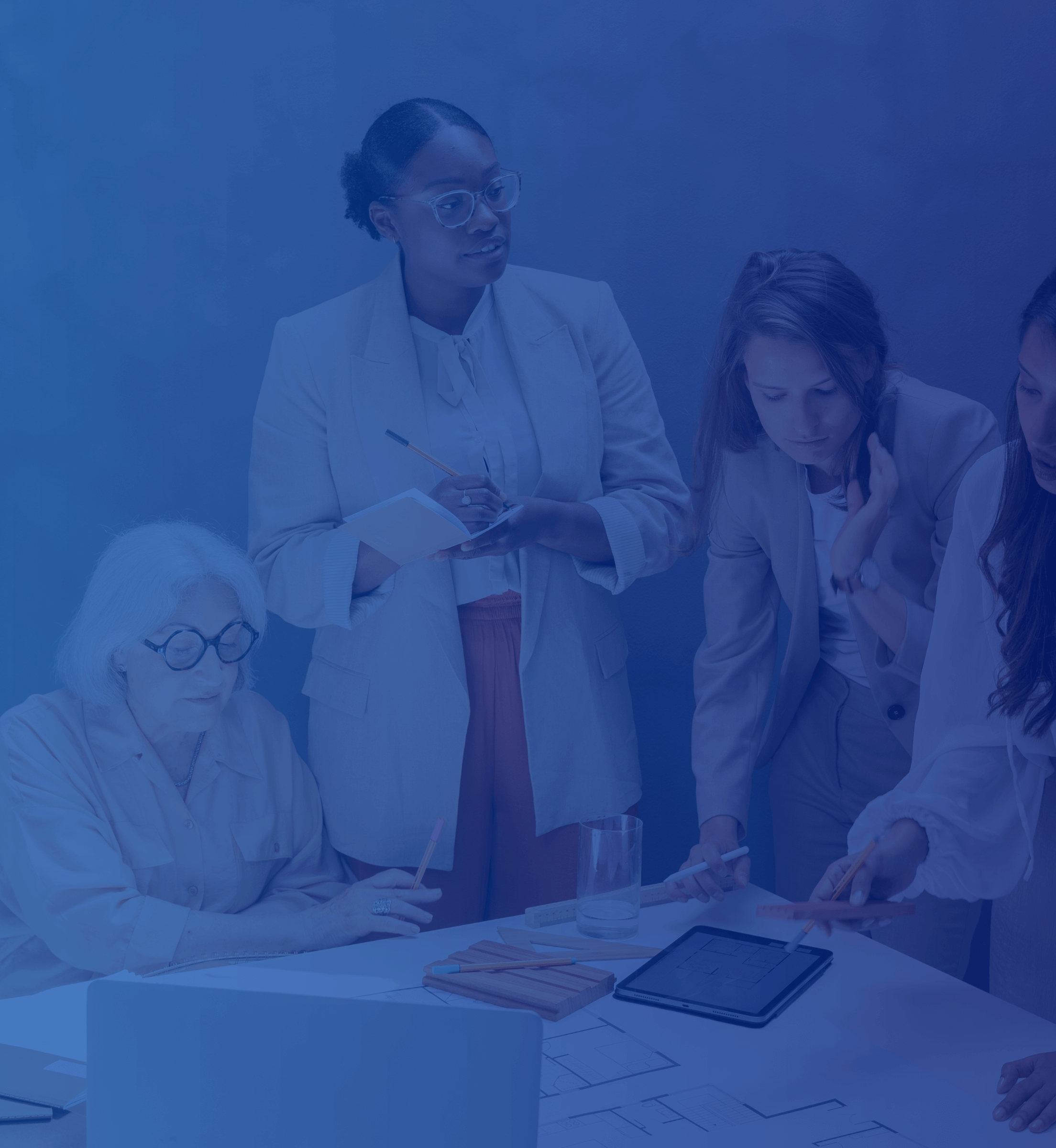 A group of diverse women around a table with notebooks and a tablet, discussing business