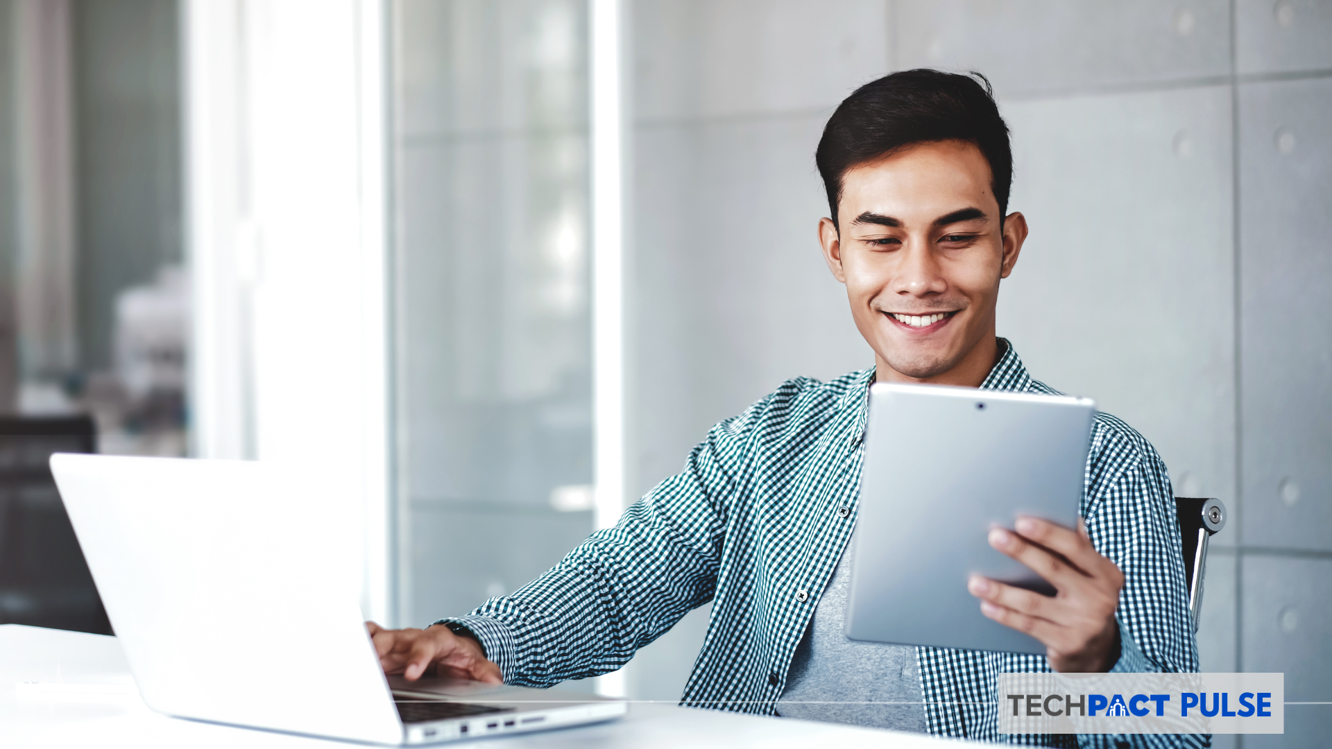 A man in an office working on a laptop while smiling on his tablet that he is holding out with one hand