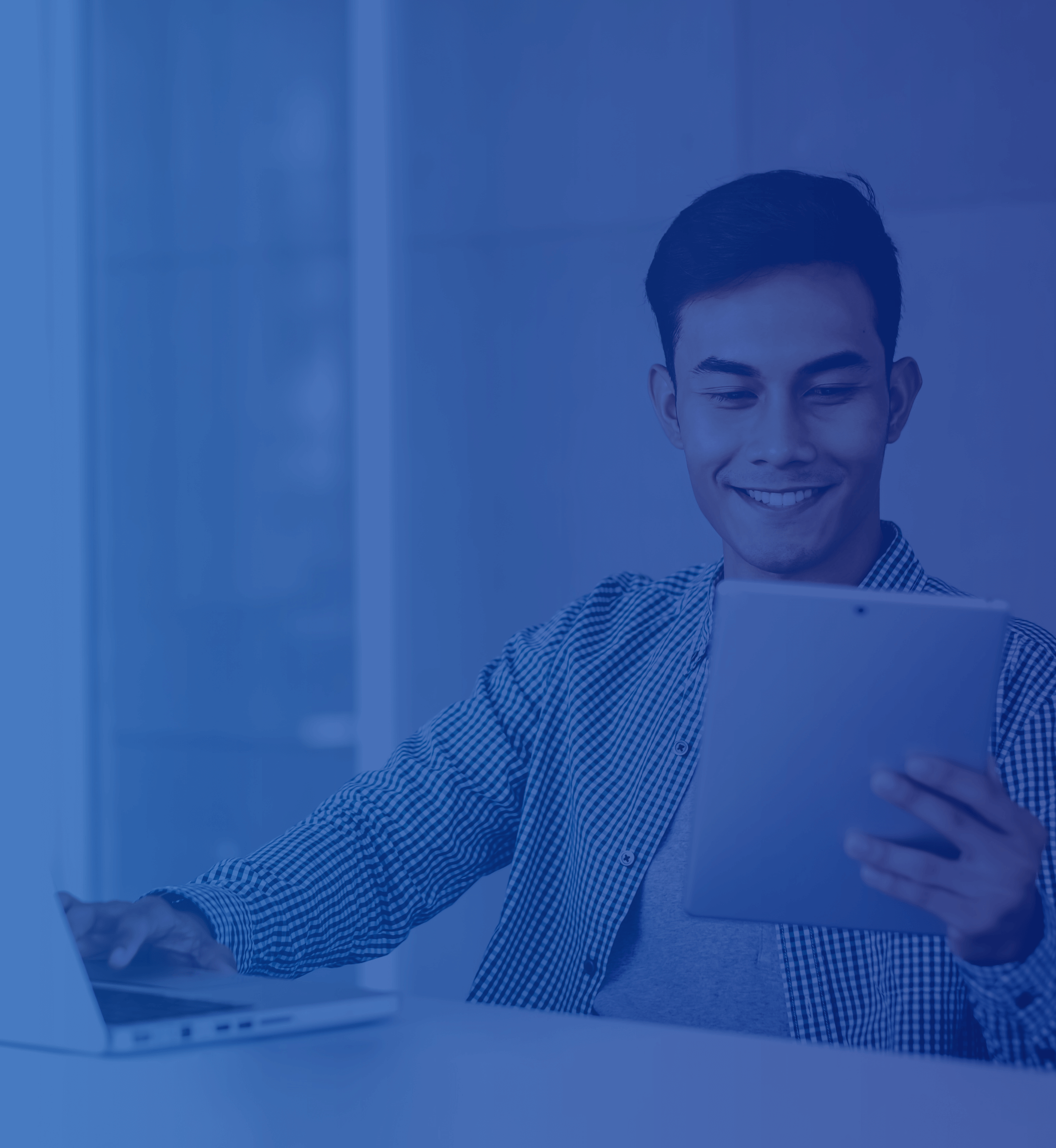 A man in an office working on a laptop while smiling on his tablet that he is holding out with one hand